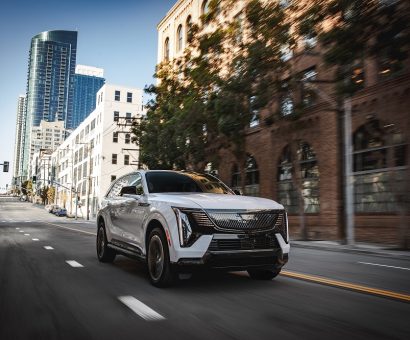 Front 3/4 view of the 2025 Cadillac ESCALADE IQ in Summit White driving on a city street.